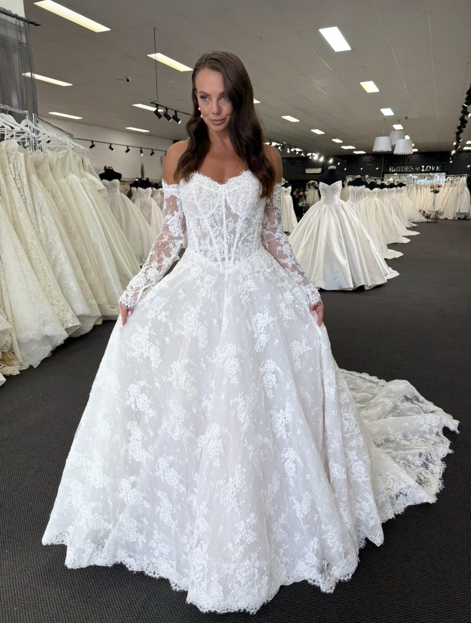 Romantic off-the-shoulder ball gown featuring a sheer illusion lace corset bodice, long fitted lace sleeves, and a full A-line skirt with delicate floral appliqué throughout. Finished with a soft lace-edged train for an elegant, timeless bridal look.