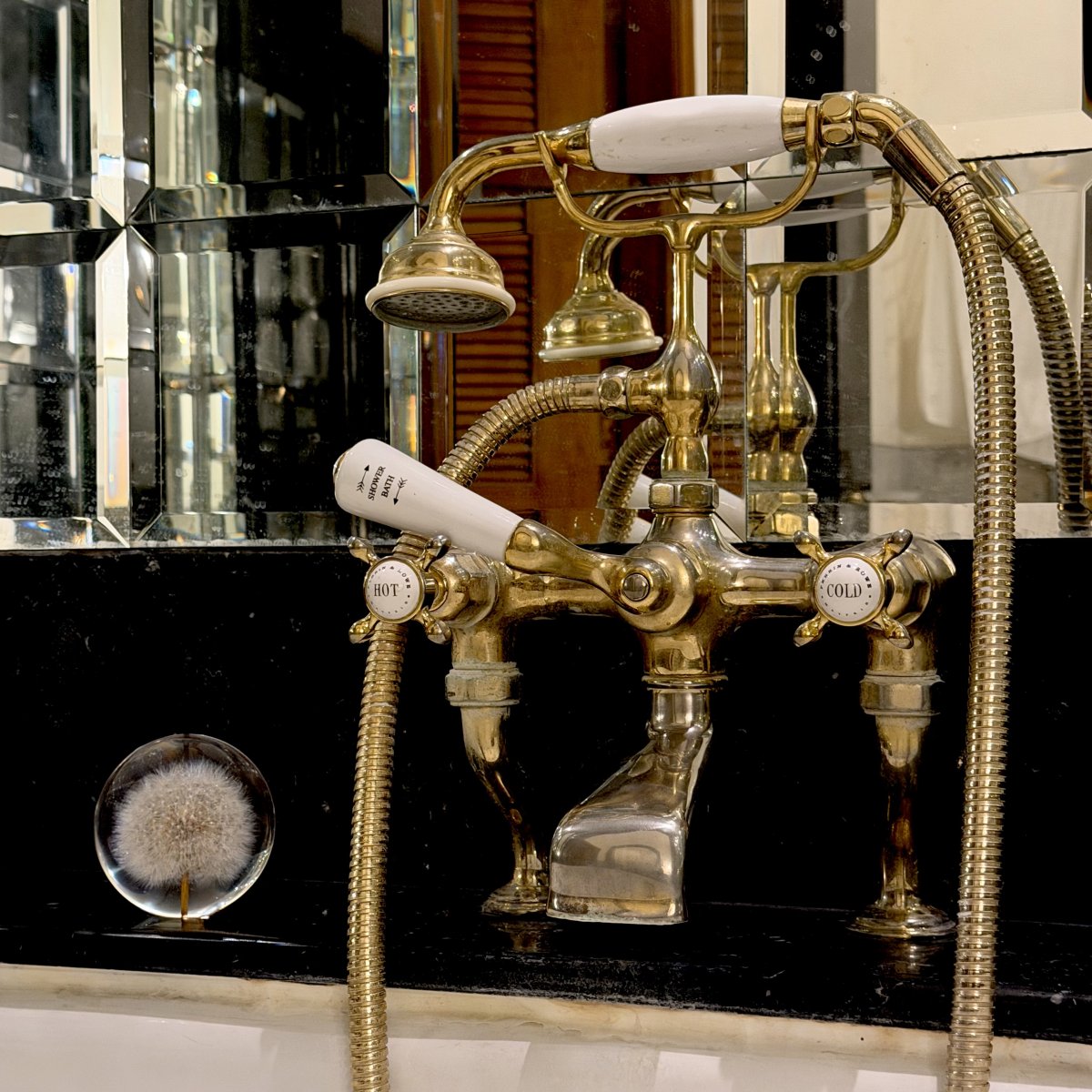 A small Dandelion Sphere with a preserved dandelion seed rests beside a polished vintage brass bath tap on a black marble ledge. The reflective mirror and classic fixtures create an elegant contrast between timeless craftsmanship and the delicate beauty of nature.