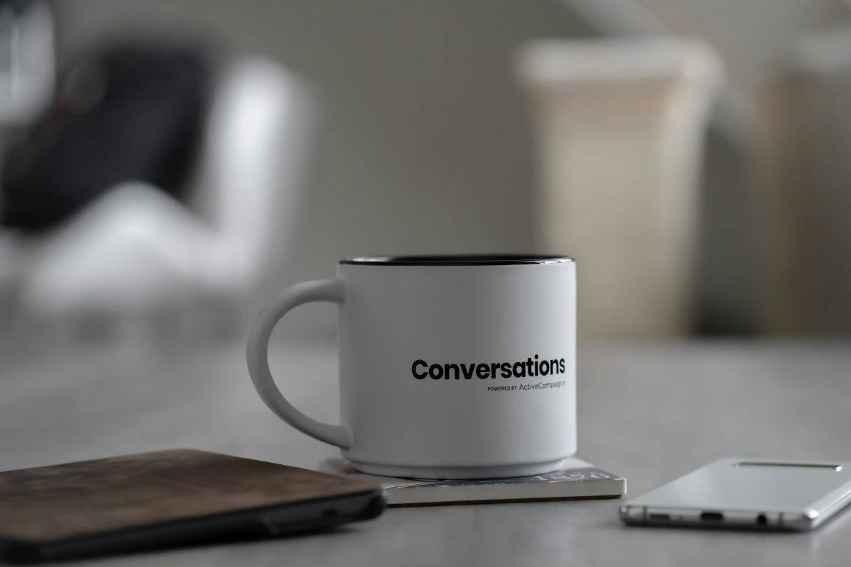 A black and white photo of a coffee mug with the word 'conversation' written on it standing on a table with a phone and a notebook alongside