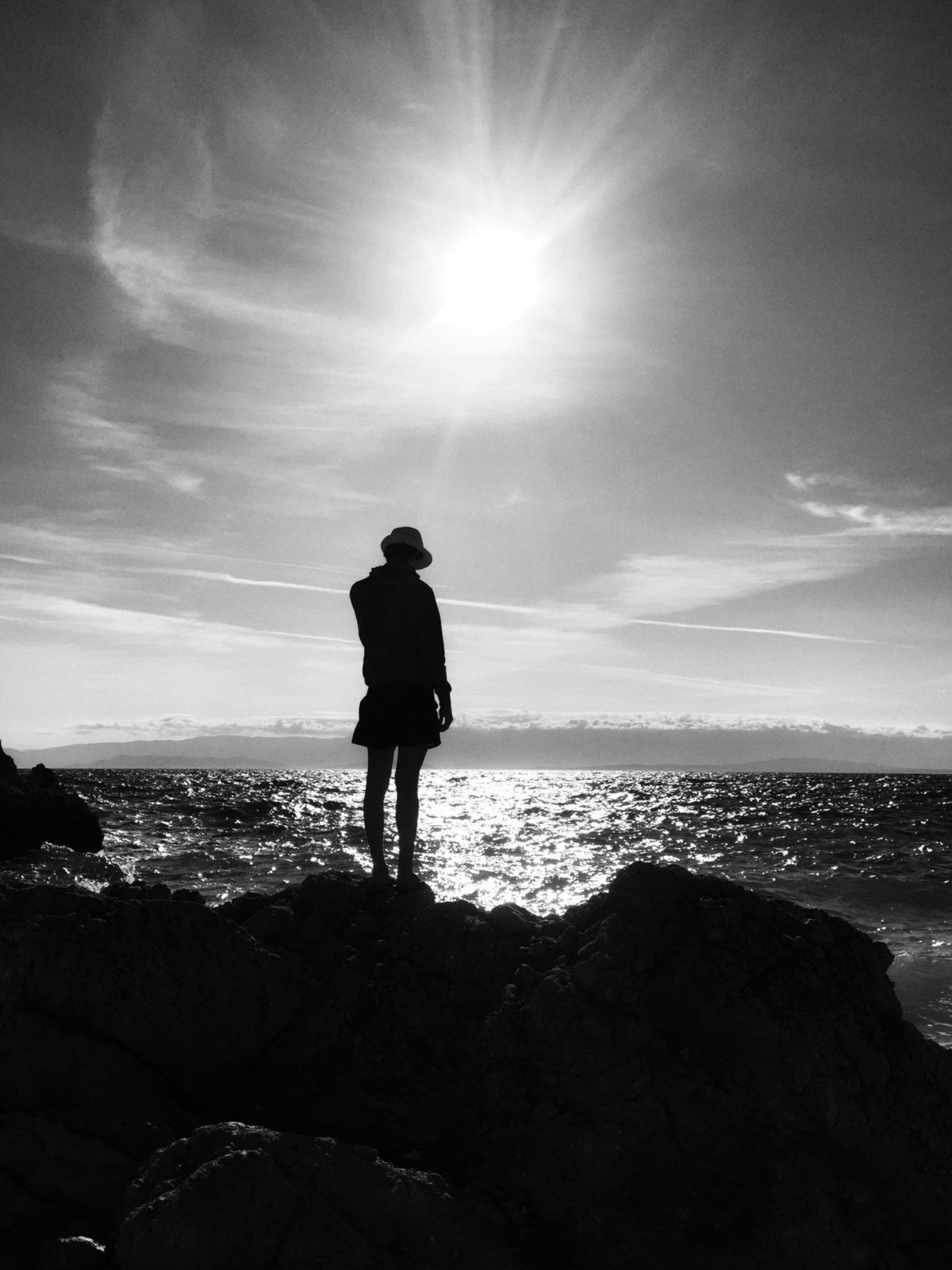 A person standing on a rocky shoreline looking out across the sea to the horizon with a sunlit route leading the way