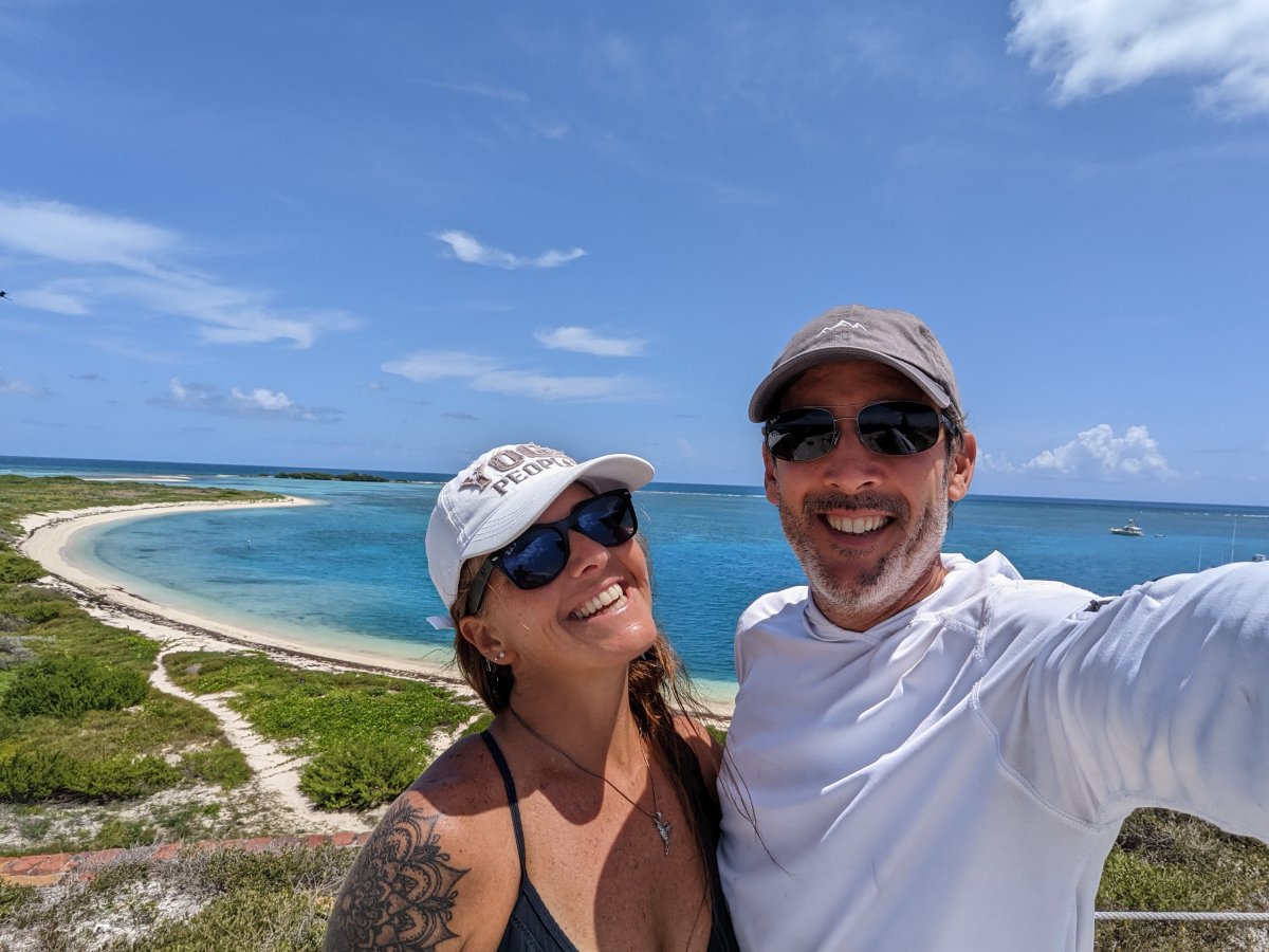 A man and woman standing together on a bright, sunny ocean beach with blue waters in the background