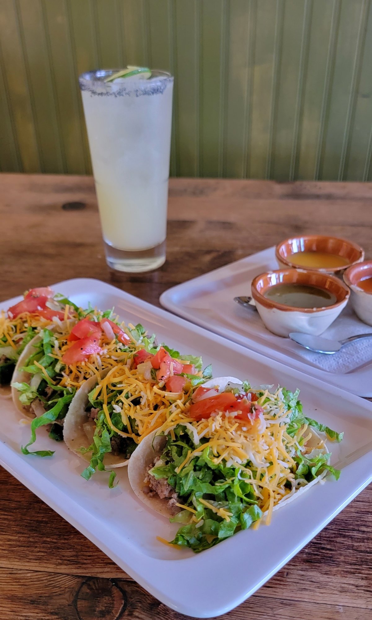 Small Bowls with brightly colored salsa.  Set of Tacos with lettuce, tomatoes, chedder-jack cheese and a margarita in the background
