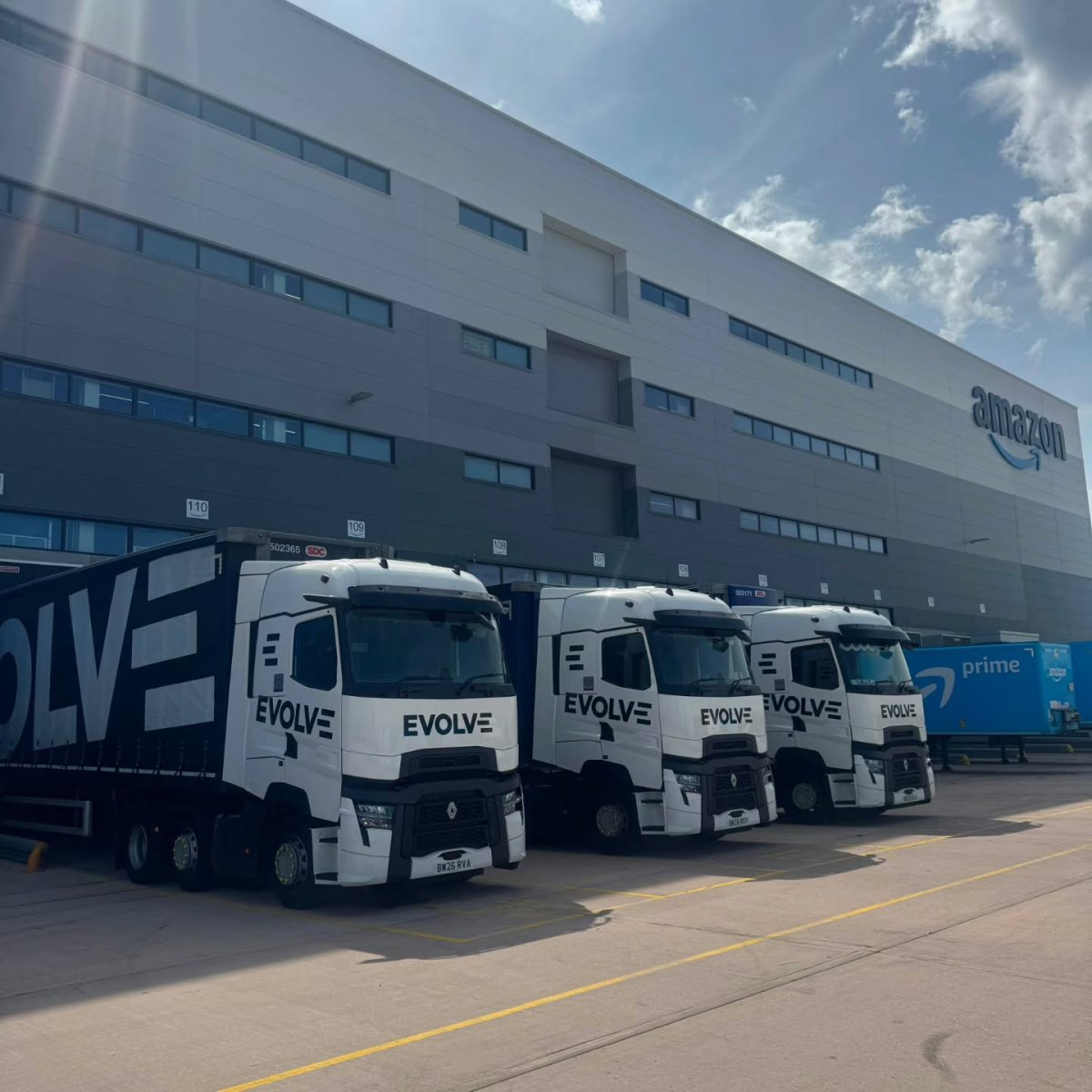 Three white Evolve tractor units with black curtain-sided trailers parked outside an Amazon distribution centre.